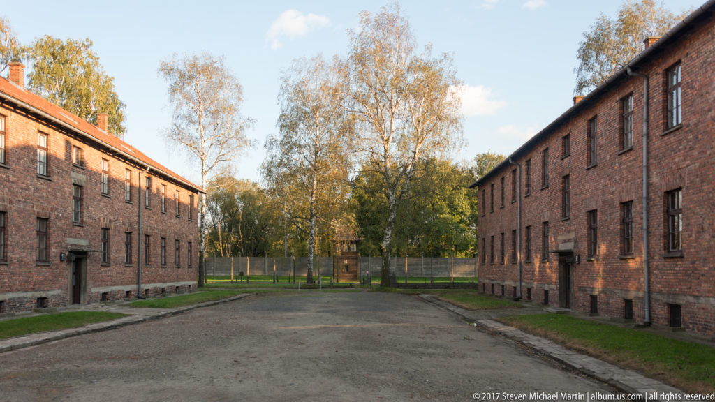 Broad yard between parallel red-brick Auschwitz I blocks, ending at trees and fencing in autumn light.
