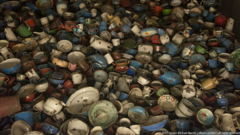 Dense pile of chipped blue, white and multicolored enamel and metal containers in an Auschwitz museum display.