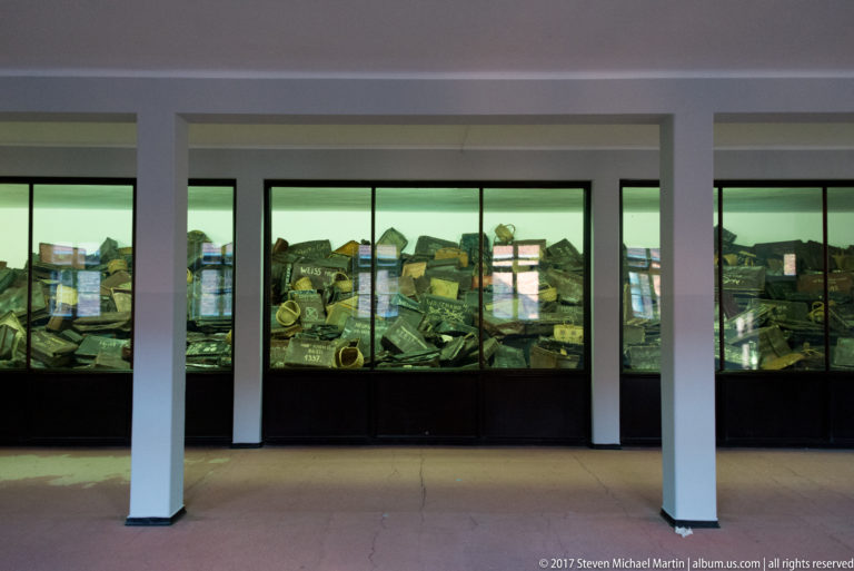 Long glass museum cases filled with confiscated belongings extend behind white columns in an Auschwitz exhibition gallery.