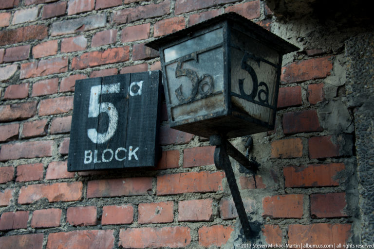 Black “5 BLOCK” sign beside an old square exterior lamp on a weathered red-brick wall.