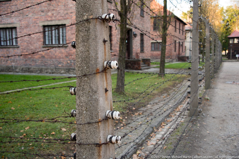 Concrete fence posts with white porcelain insulators and barbed wire recede beside red-brick Auschwitz I blocks.
