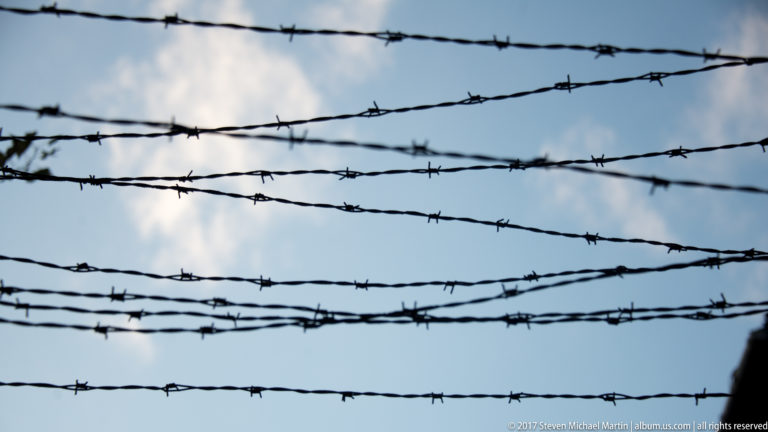 Multiple horizontal strands of dark barbed wire silhouetted against a pale blue, clouded sky.