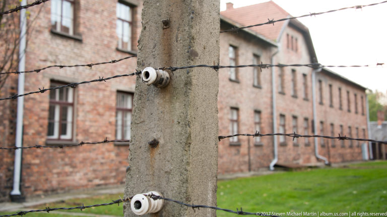 Concrete fence post with white porcelain insulators and barbed wire, with a red-brick camp block blurred behind.