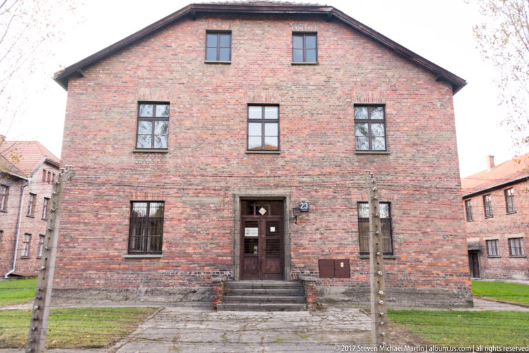 Front of red-brick Auschwitz I Block 23 with central wooden door, steps, windows and perimeter posts.