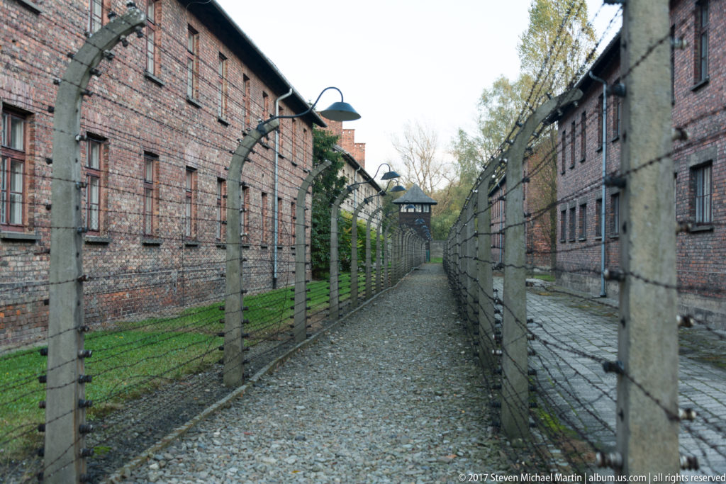 Gravel corridor between parallel barbed-wire fences and brick blocks, ending at a distant guard tower.
