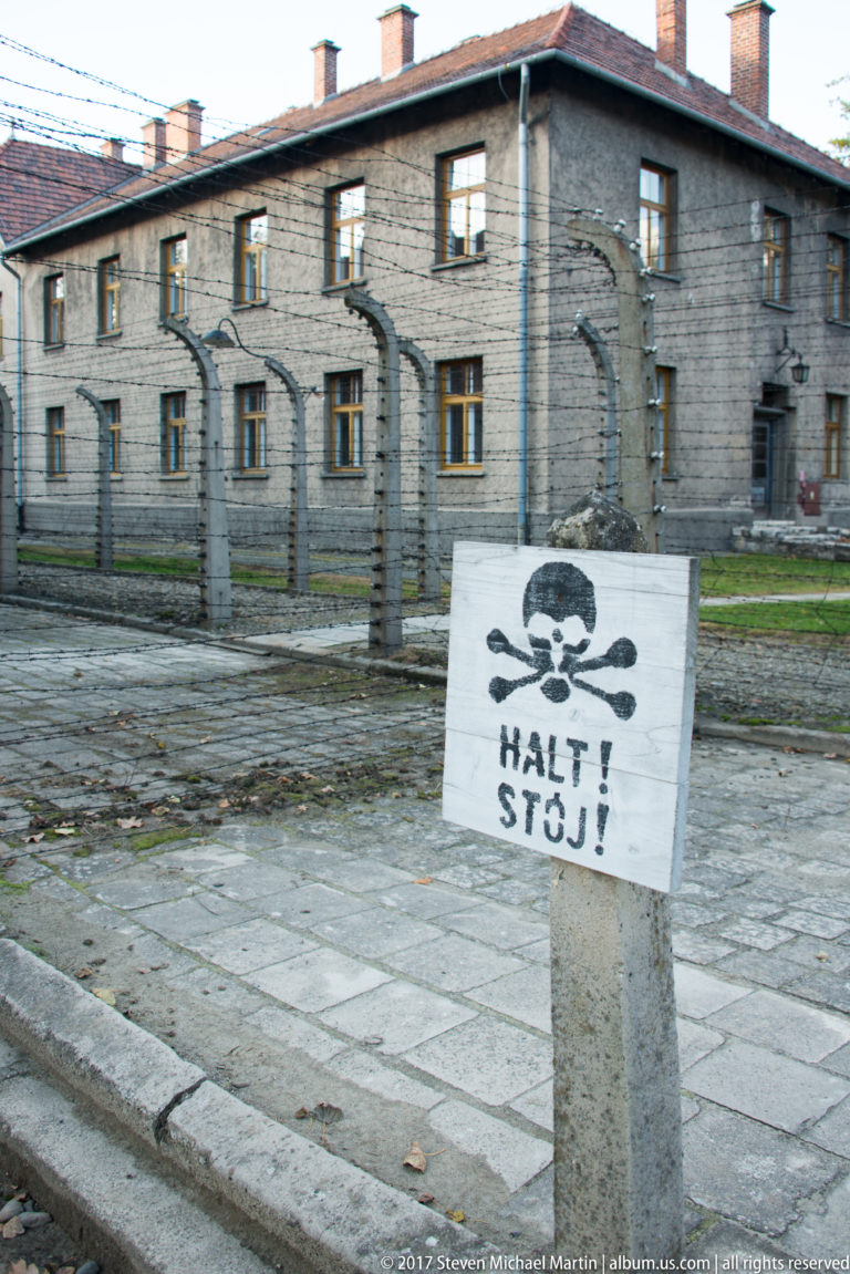White skull-and-crossbones sign reading “HALT! STÓJ!” on a concrete post beside the Auschwitz I fence.