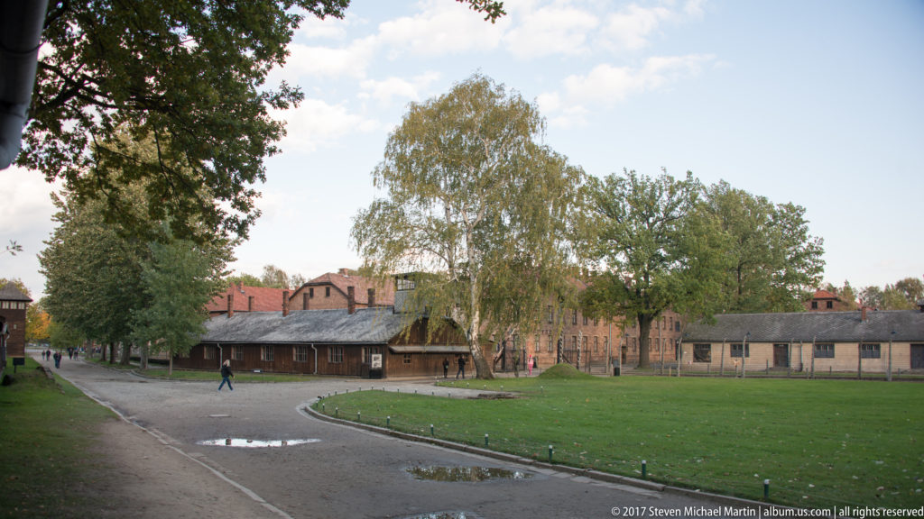 Curving road and lawn lead past low camp buildings and mature trees toward larger brick blocks at Auschwitz I.