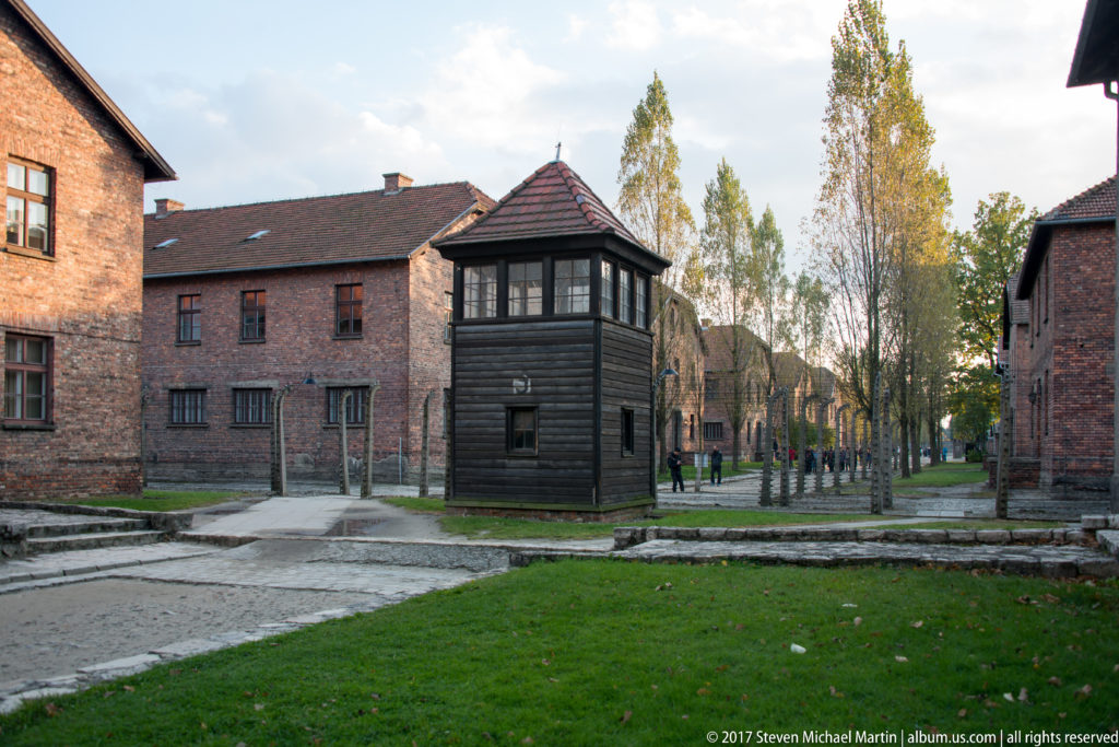 Dark wooden guard tower stands among red-brick camp blocks, perimeter fencing, lawn and autumn trees.