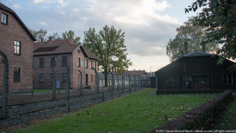 Electrified fence recedes between red-brick blocks and a dark wooden building across grassy Auschwitz I grounds.