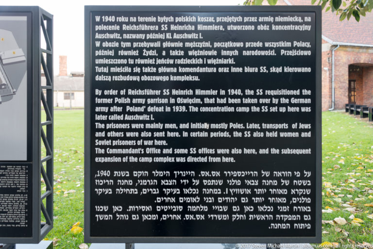 Multilingual Auschwitz historical panel in Polish, English and Hebrew with camp buildings visible behind it.