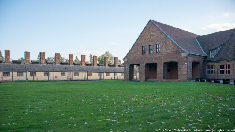 Green lawn fronts a red-brick gateway-like building and a long pale barrack with many chimneys.