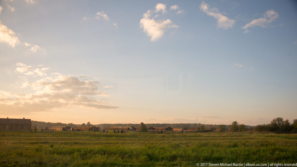 Open grassy grounds at Auschwitz II-Birkenau with low barracks and camp structures stretching across the distant horizon.