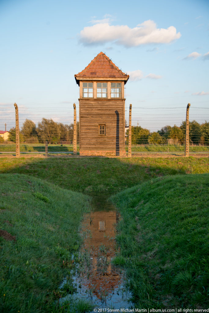 Centered wooden guard tower behind barbed-wire fencing and wet grass at Auschwitz II-Birkenau.