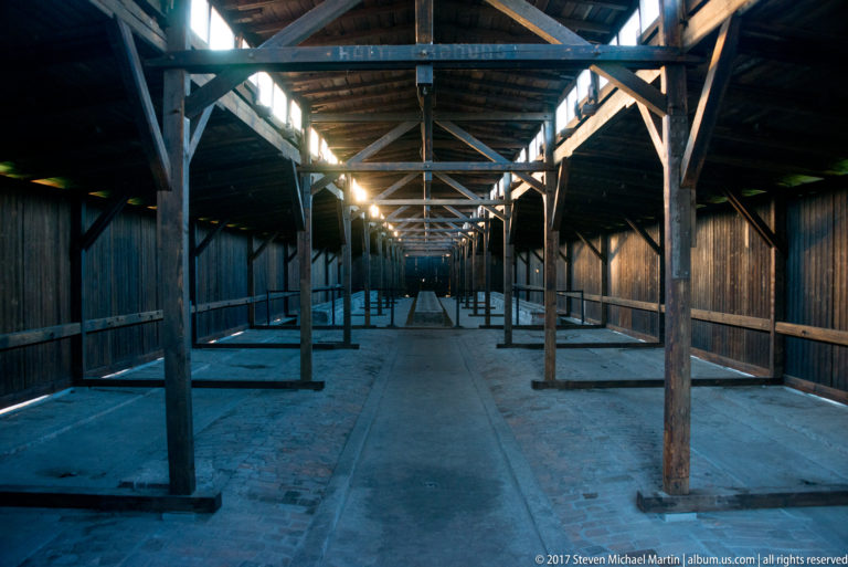 Interior of a wooden barrack with exposed trusses, rows of posts, and a central brick-paved aisle.