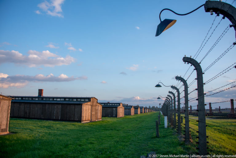 Curved concrete fence posts with barbed wire and lamps beside wooden barracks at Auschwitz II-Birkenau.