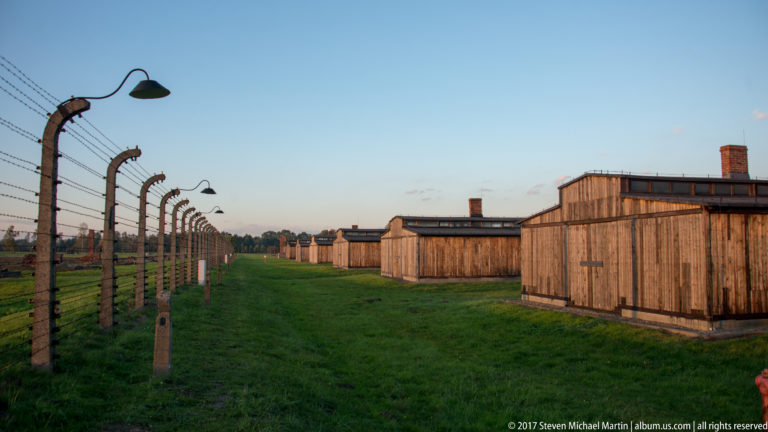 Long barbed-wire fence with concrete posts and lamps running beside rows of wooden barracks.