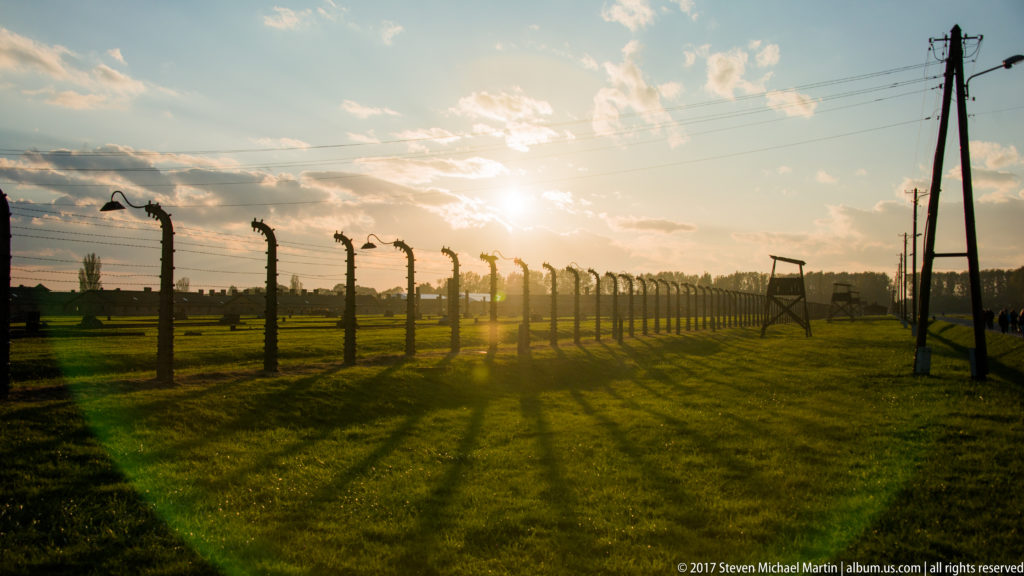 Barbed-wire perimeter fence and wooden guard tower at Auschwitz II-Birkenau backlit by a low evening sun.