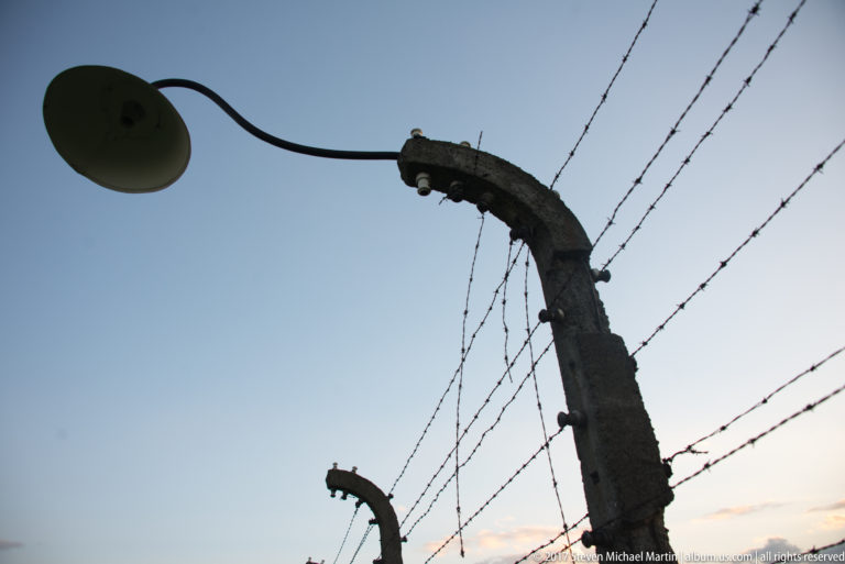 Upward view of a curved concrete fence post, barbed wire, and lamp fixture at Auschwitz II-Birkenau.