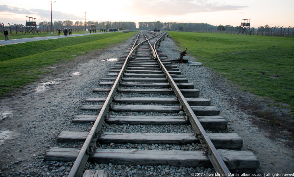 Branching railway tracks at Auschwitz II-Birkenau with perimeter fencing and a distant guard tower.