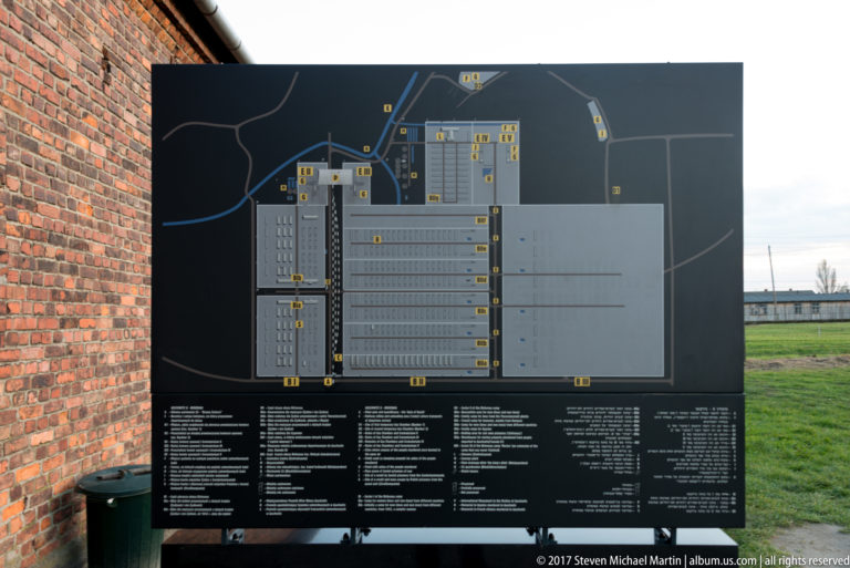 Large site-plan information panel for Auschwitz II-Birkenau beside a brick wall, with camp grounds visible beyond.