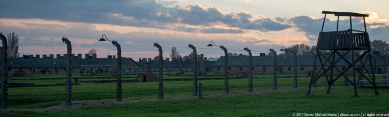 Wooden guard tower beside barbed-wire fencing, with brick barracks and chimneys beyond at Auschwitz II-Birkenau.