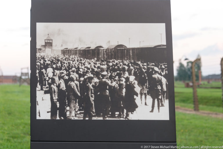 Interpretive panel displaying a historical crowd beside railway cars, with the Birkenau grounds blurred behind it.
