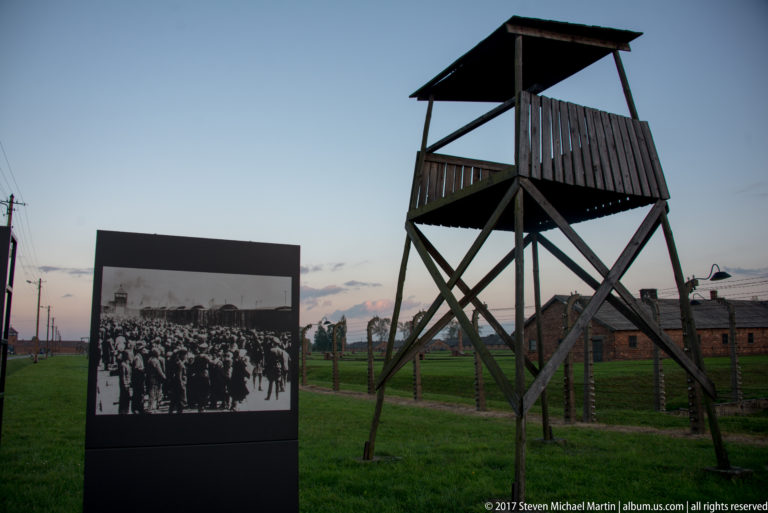 Wooden guard tower beside a historical photograph panel, with fencing and brick camp buildings in the background.