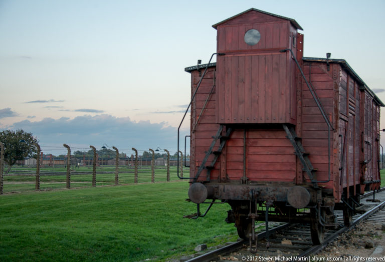 End view of a weathered red wooden freight wagon on rails beside the fenced grounds at Auschwitz II-Birkenau.