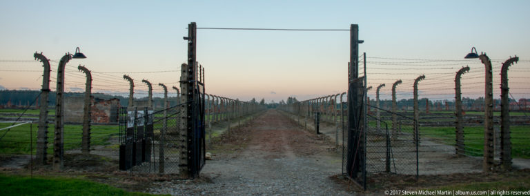 Open metal gate in the barbed-wire perimeter, framing a straight gravel path between parallel fences at Auschwitz II-Birkenau.