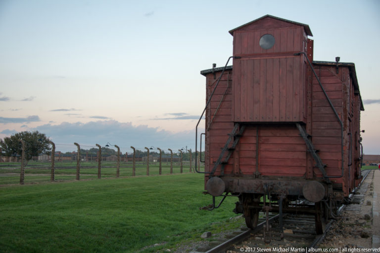 Centered rear view of a weathered red wooden freight wagon with buffers and fencing behind it at Auschwitz II-Birkenau.