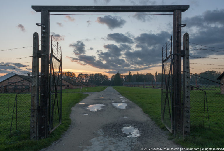 Open fenced gateway framing a puddled paved road and low camp buildings at dusk at Auschwitz II-Birkenau.