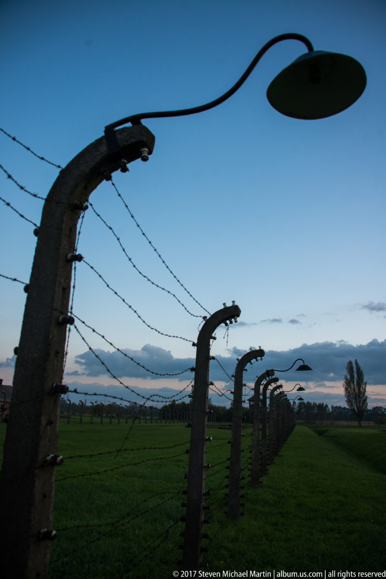 Curved concrete fence posts with barbed wire and an oval lamp receding beneath a blue evening sky.