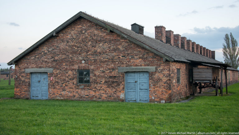 Brick barrack with two pale blue doors, a central window, pitched roof, and multiple chimneys at Auschwitz II-Birkenau.