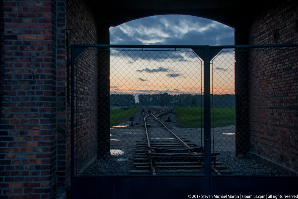 Railway tracks seen through the brick entrance arch and metal gate of Auschwitz II-Birkenau at dusk.