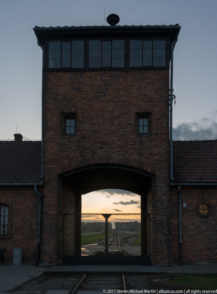 Frontal view of the brick Auschwitz II-Birkenau entrance gatehouse with railway tracks passing through its central arch.