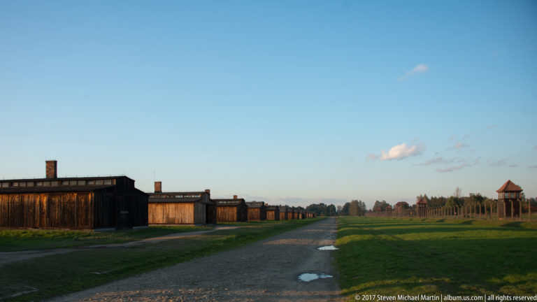Gravel road with puddles between wooden barracks, fencing, and distant guard towers at Auschwitz II-Birkenau.