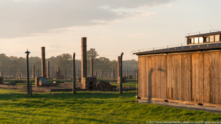 Brick chimneys and low barrack ruins across grass beside a surviving wooden barrack at Auschwitz II-Birkenau.