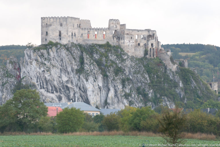 Side view of Beckov Castle ruins stretched along a sheer limestone crag above trees and farmland.