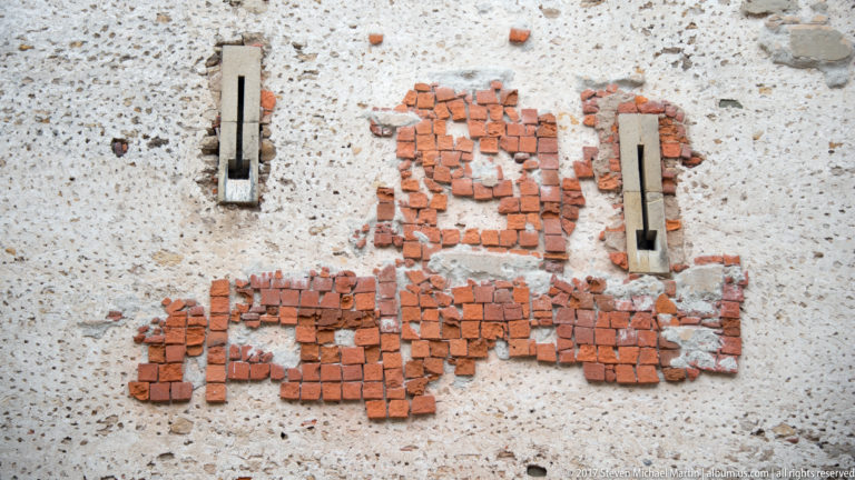 Red brick patches and two narrow openings exposed within a pale rendered historic wall.