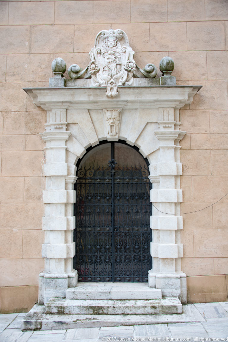 Decorated arched doorway with pale stone surround, heraldic crest, and dark metal grille in a warm ashlar wall.