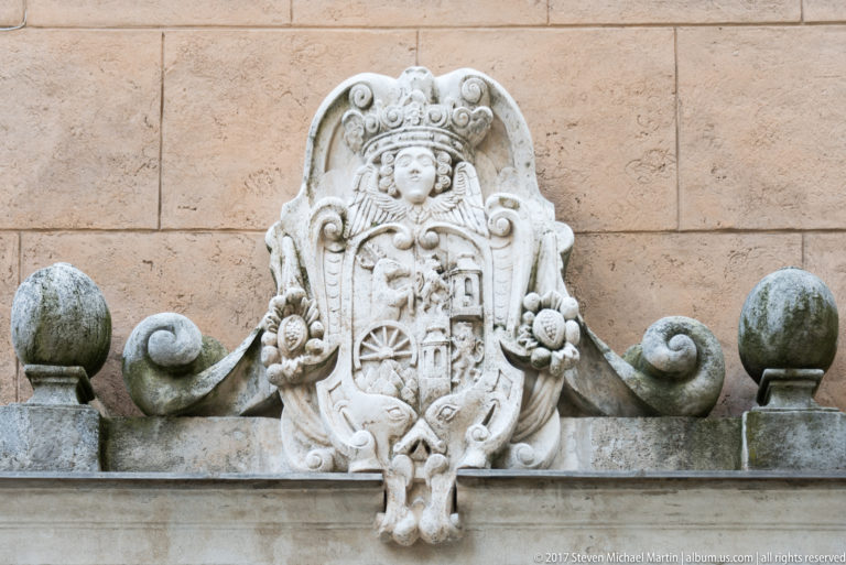 Weathered pale stone heraldic cartouche with crown, scrolls, and relief symbols above a castle entrance.
