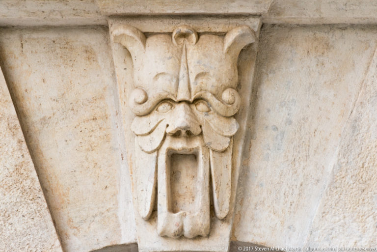 Close view of a weathered stone grotesque mask with curled moustache and open mouth.