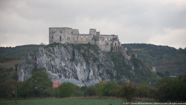 Distant profile of Beckov Castle ruins atop a high limestone rock above open fields and trees.