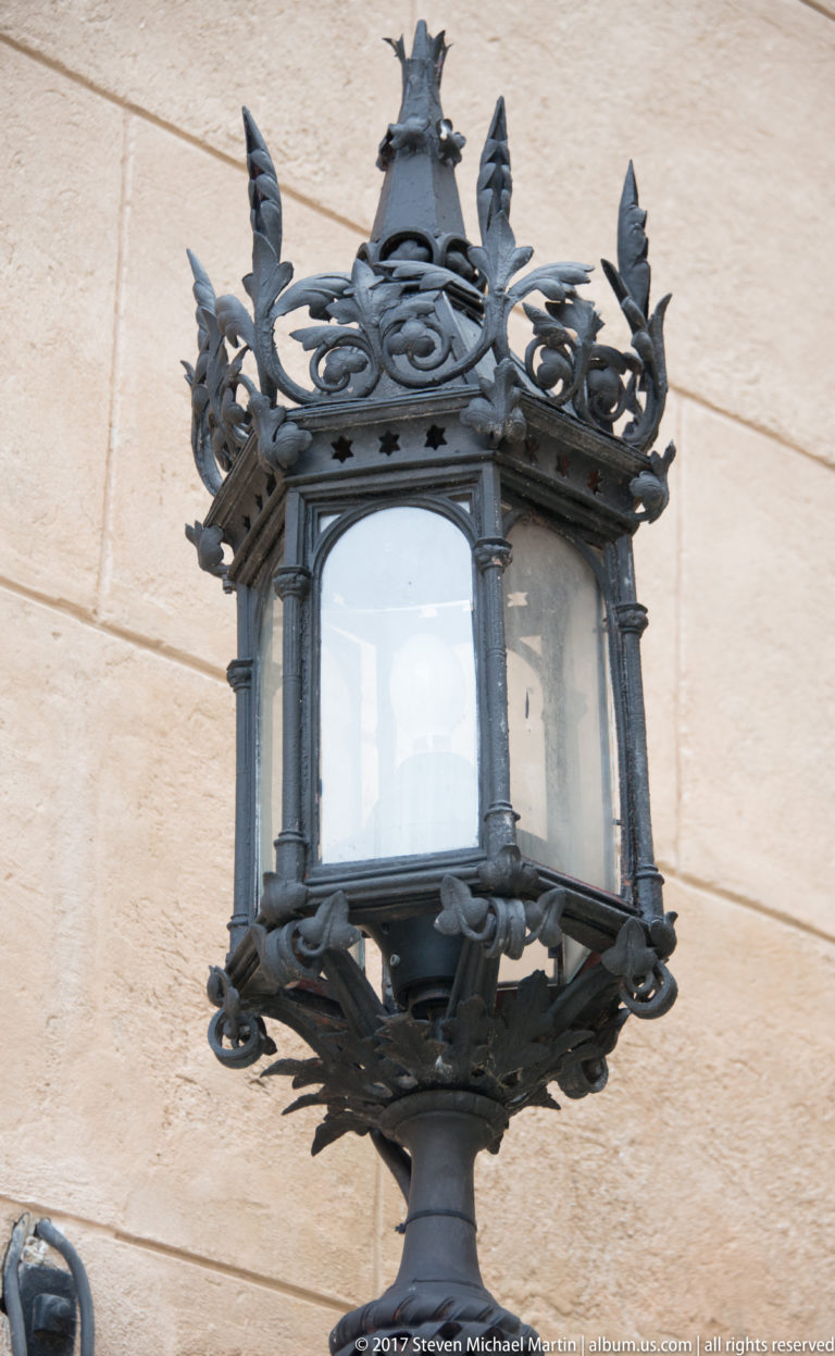 Ornate dark metal wall lantern with glazed panels, scrollwork, and leaf-shaped finials mounted on pale stone.