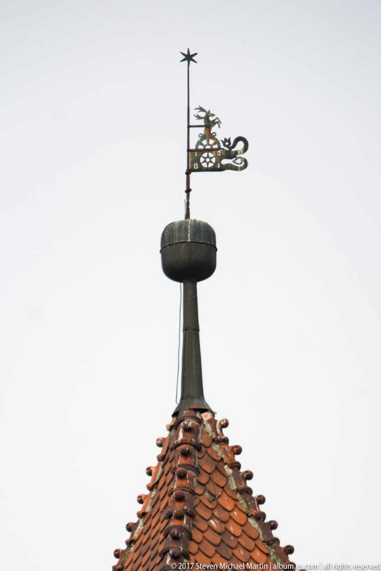 Slender decorative weather vane and openwork finial rising above a red-tiled castle roof.