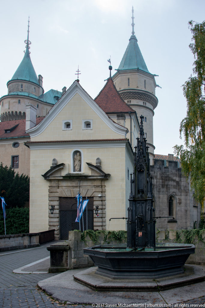 Bojnice Castle forecourt with pale entrance building, ornate dark fountain, flags, and round towers behind.