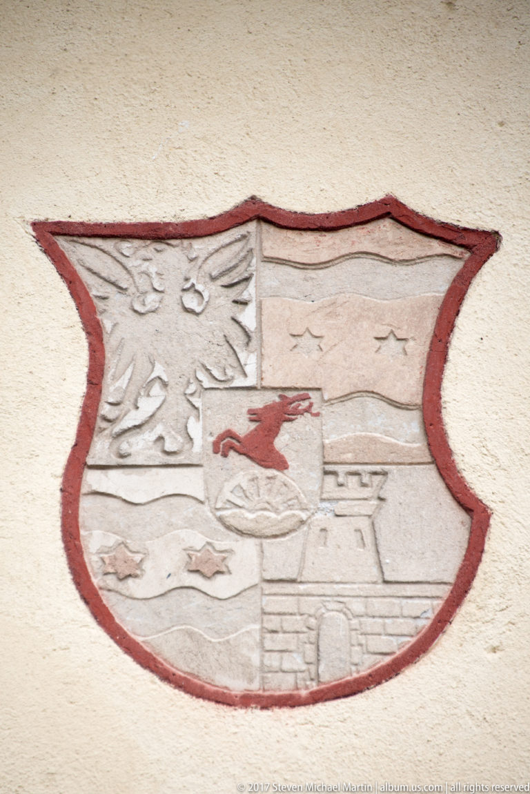 Painted shield-shaped heraldic emblem with birds, red animal, crescent motif, and stylized gateway on plaster.