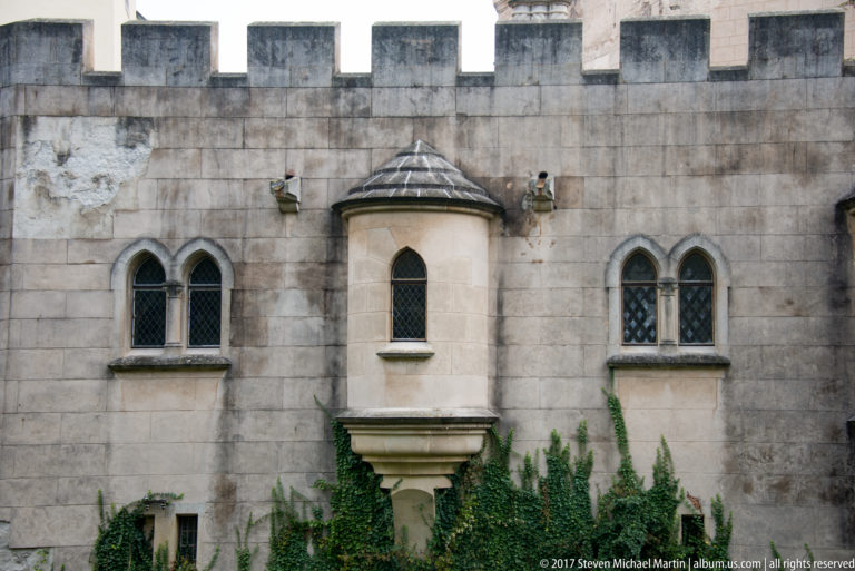 Crenellated castle wall with projecting stone oriel windows, pointed openings, and climbing ivy.