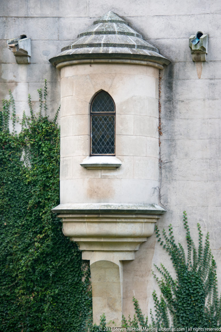 Small projecting polygonal window bay with arched glazing, faceted roof, stone corbel, and ivy.