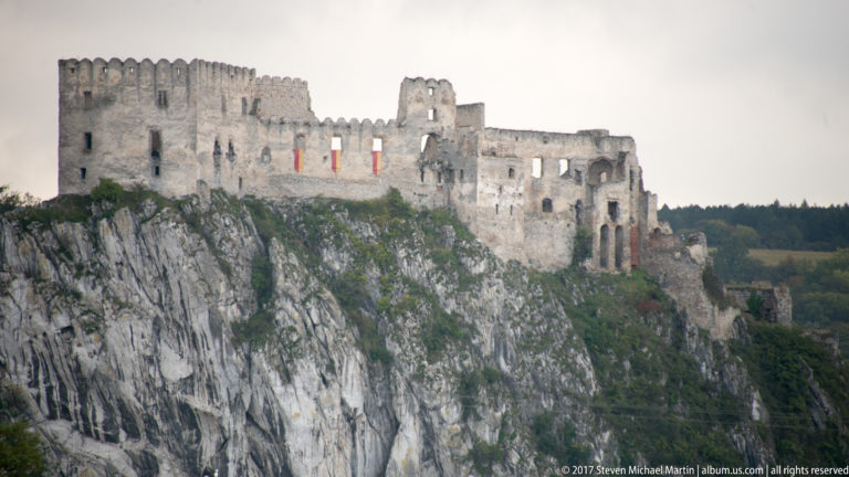Telephoto view of Beckov Castle’s roofless walls and window openings above a striated limestone cliff.
